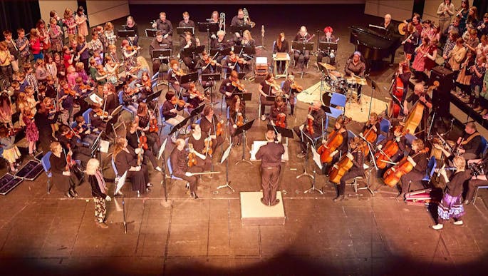 Seen from above, an orchestra made up of adults and children perform together on stage.