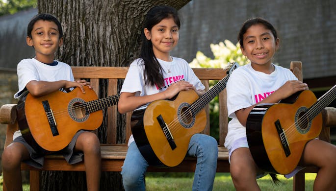 Three kids, one boy and two girls, hold guitars and smile, sitting on a bench in front of a tree.