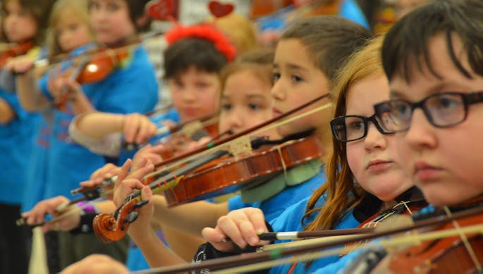 Young children play violins in a classroom, all wearing Juneau Alaska Music Matters blue t-shirts.