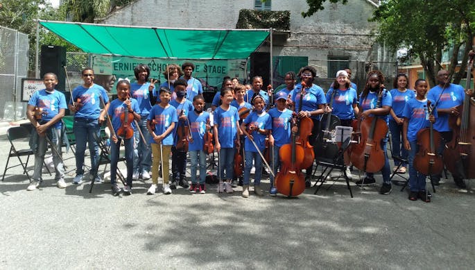A group of kids stand outside with their string instruments, all wearing Make Music NOLA t-shirts.
