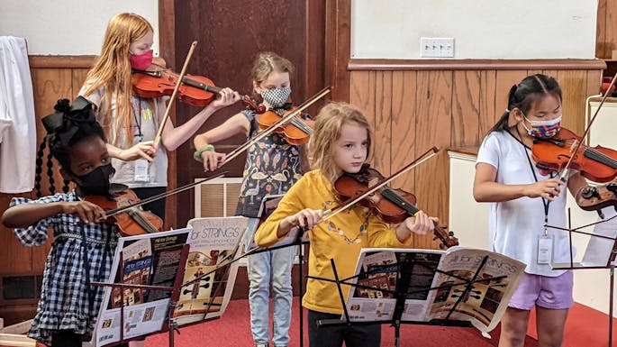 Children stand in front of music stands and play violin together.