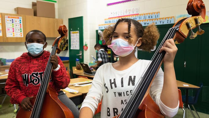 In a classroom, two kids wearing masks stand and pose with their cellos.