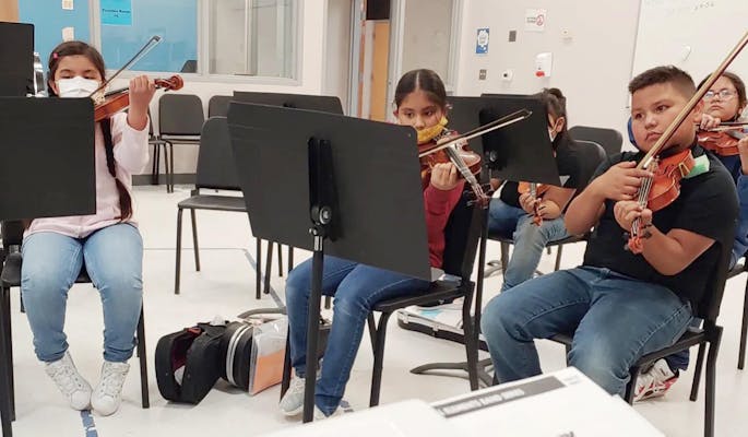 Children practice violin together in a classroom filled with music stands.