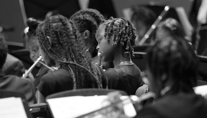 In black and white, a group of young people play music in rehearsal. A single girl is focused on.
