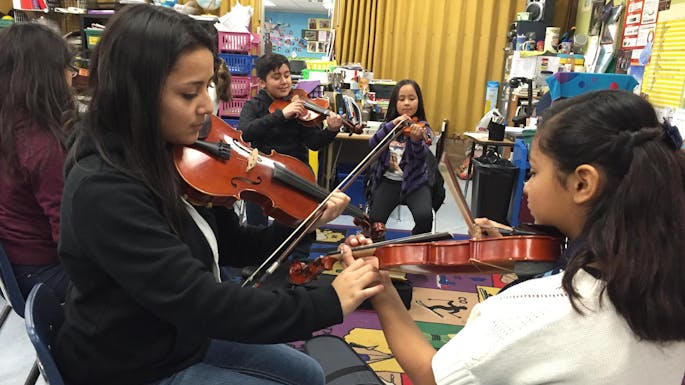 Five girls play cello together in rehearsal. Music stands fill the room.