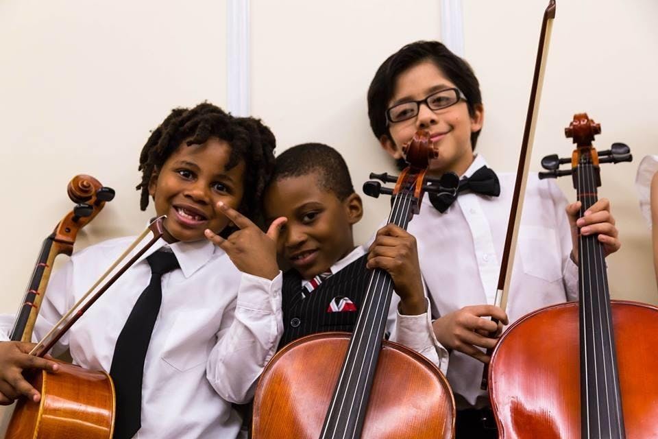 Three young boys in formal attire hold cellos and smile against at white wall.