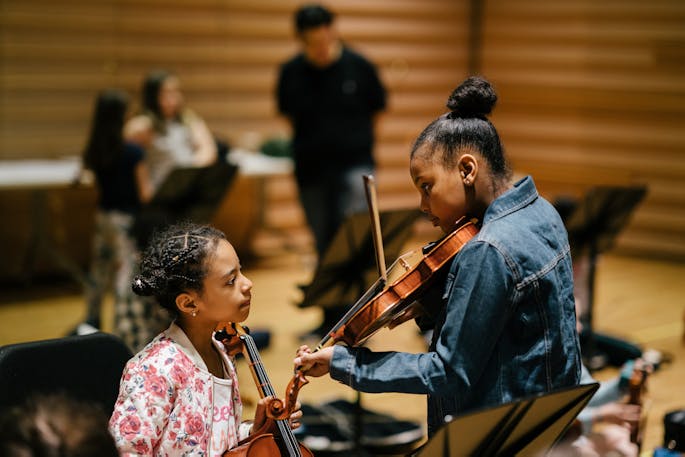 An older girl in a jean jacket shows a younger girl in a floral jacket her violin in a rehearsal.