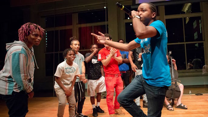 A teen boy sings into a microphone as other teens cheer him on.