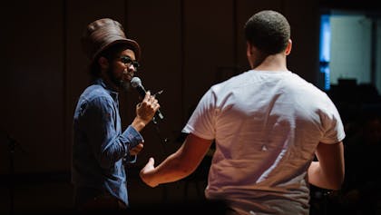 A young man holding a microphone talks with another young man.