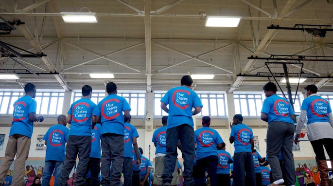A group of young men wearing blue "Our Voices Count" t-shirts stand on a riser before microphones