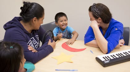 Two female teachers sit at a table with a smiling young boy as he colors.