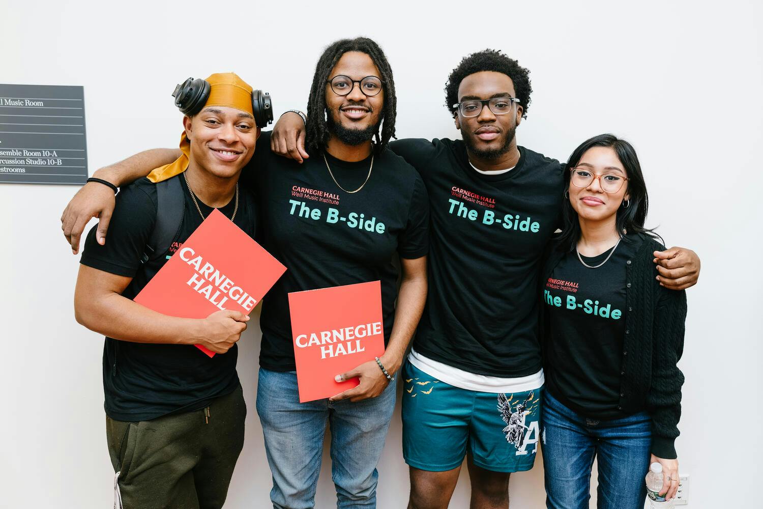 Three young men and a young woman pose with their arms around each other, wearing black "The B-Side" t-shirts