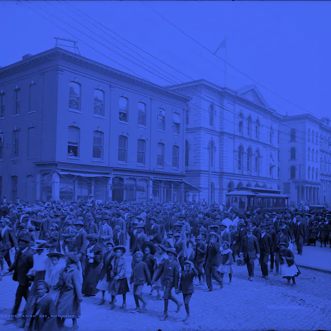 A blue-hued photo of a crowd of mostly black men, women, and children crossing the street before a row of buildings in 1865