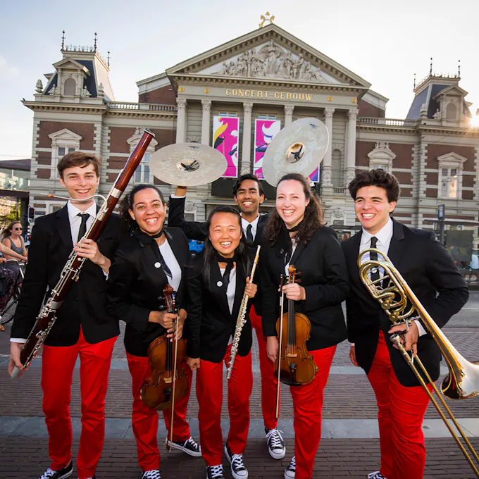 Young musicians in black blazers and red pants stand holding their instruments outside the concert hall in Amsterdam
