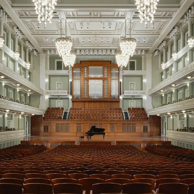 A concert hall with white box seats, chandeliers hanging, and a grand piano on stage