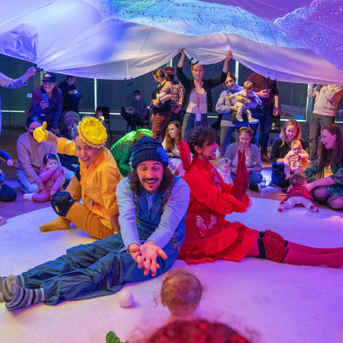 Performers dressed in colorful outfits sit on the ground interacting with toddlers
