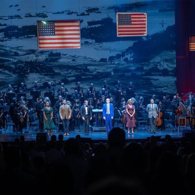 Performers and an orchestra on stage, with American flags and a projected image backdrop