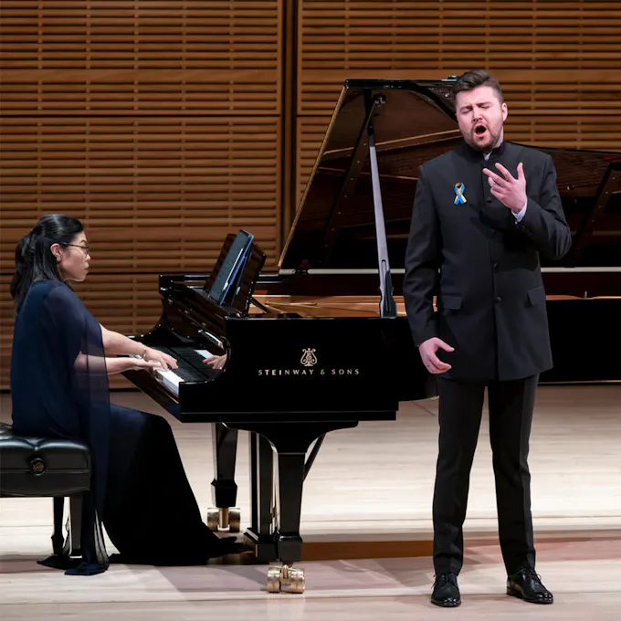 A man sings in front of a piano as a woman plays