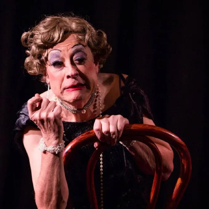 A man dressed in drag, make-up, and jewelry sits on a chair, hand raised