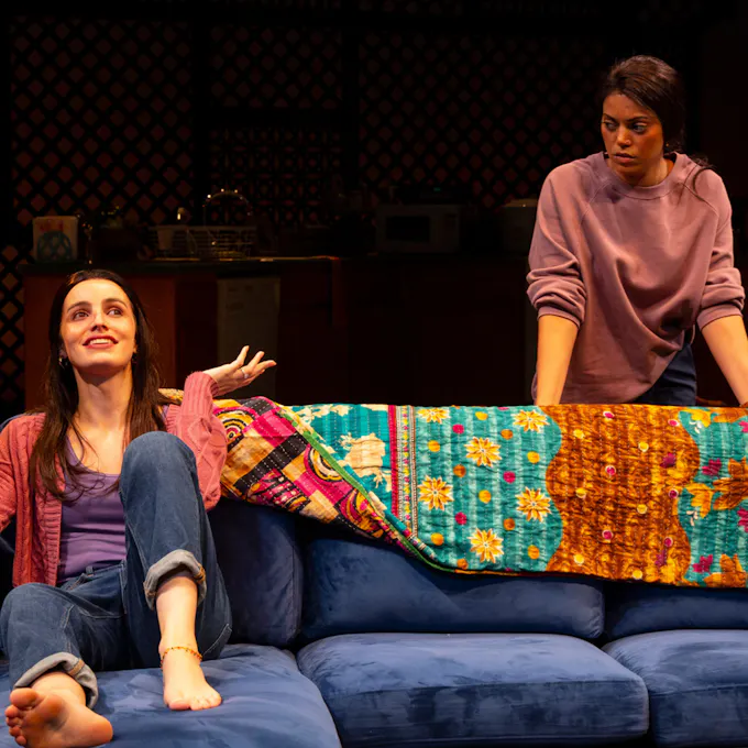 A woman sits on a couch smiling as another woman looks on
