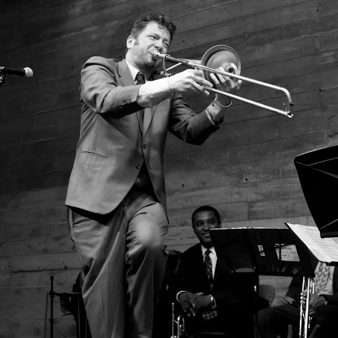 A man in a suit plays trombone, one leg raised, as two seated trumpeters watch