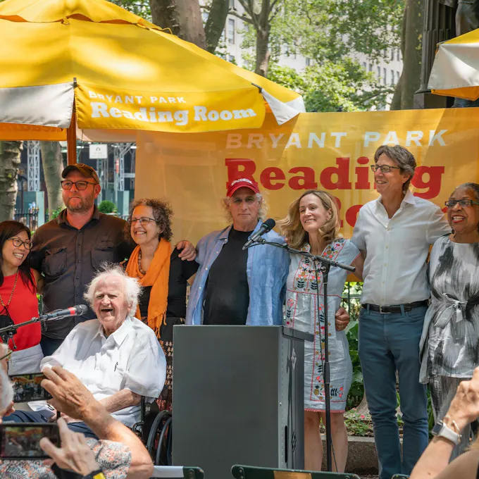 A group of people outdoors, arms around each other, before a banner: "Bryant Park Reading Room"