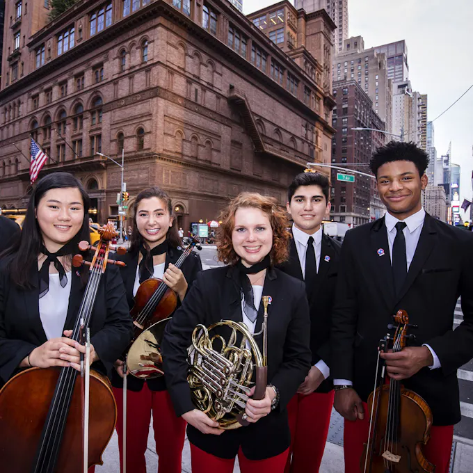 Five teenage musicians wearing black blazers and red pants stand on the corner outside Carnegie Hall.