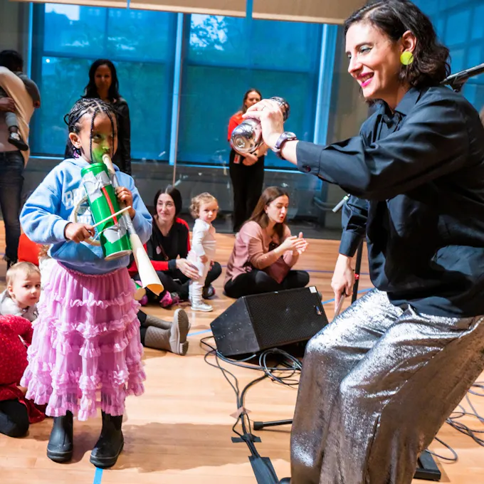 Five musicians in colorful clothing perform for an audience of children playing homemade instruments