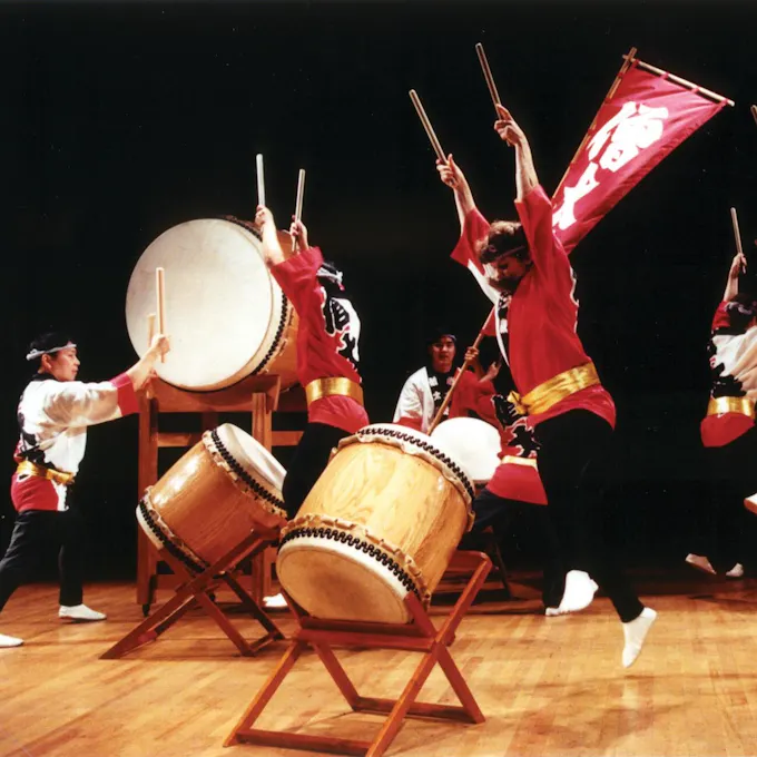 Performers wearing red jackets leap into the air holding drumsticks, preparing to beat their drums on stage