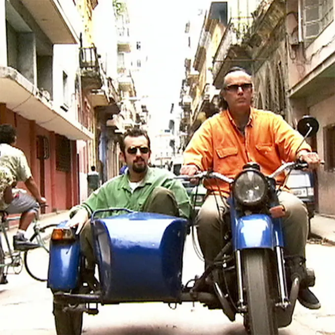 Two men on a blue motorcycle and sidecar ride down a village street