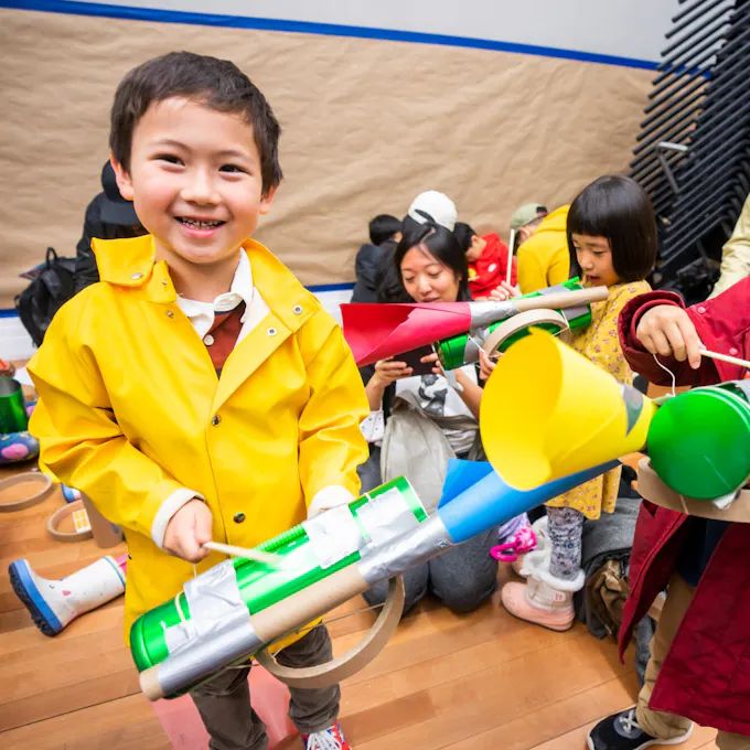 Five musicians in colorful clothing perform for an audience of children playing homemade instruments