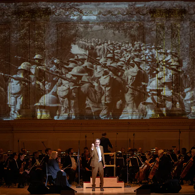 A performer standing and speaking on stage, with a hand raised. An orchestra behind, with a video projection above of soldiers walking down a road