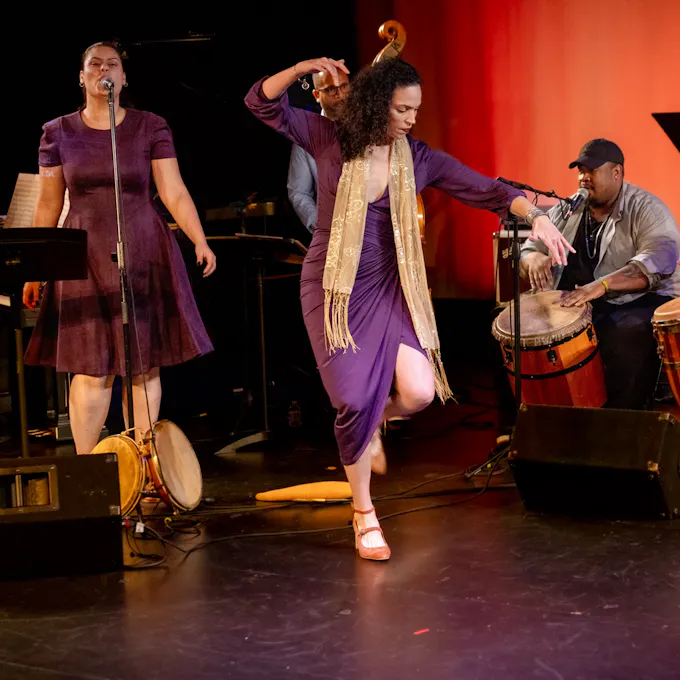 A young woman wearing a purple dress and gold scarf dances as another sings and a man plays conga