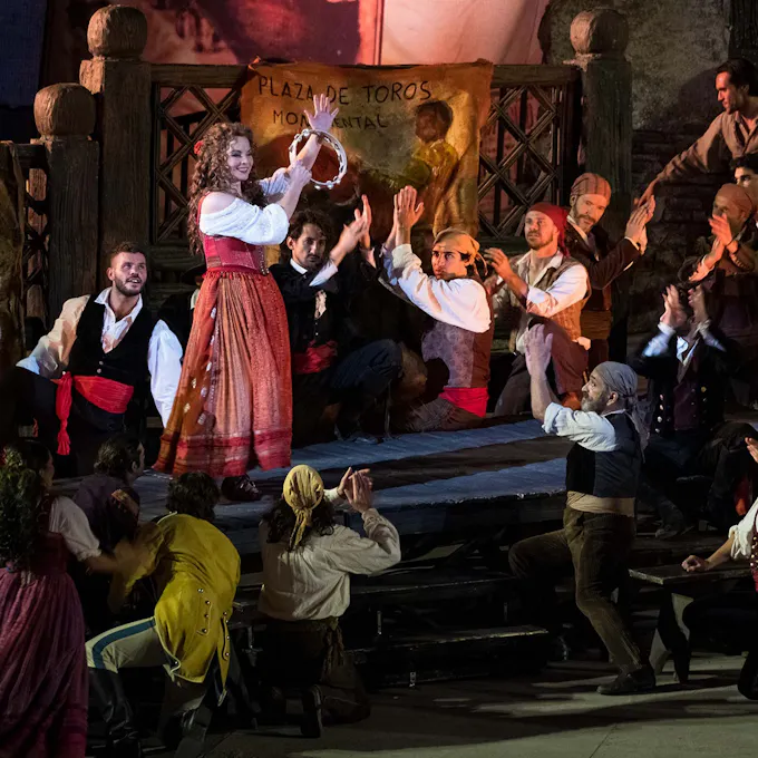 An actor in a red dress plays tambourine as performers stand around clapping