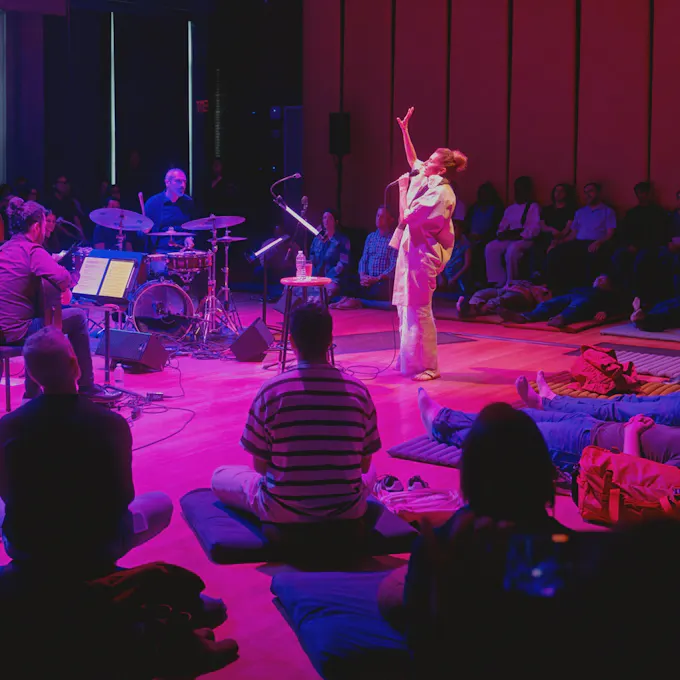 A woman sings, hand uplifted, as people sit and lie on mats around her