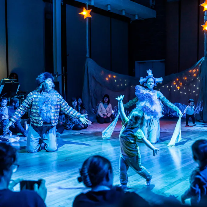 Performers in costume kneel on a stage lighted blue as a child dances
