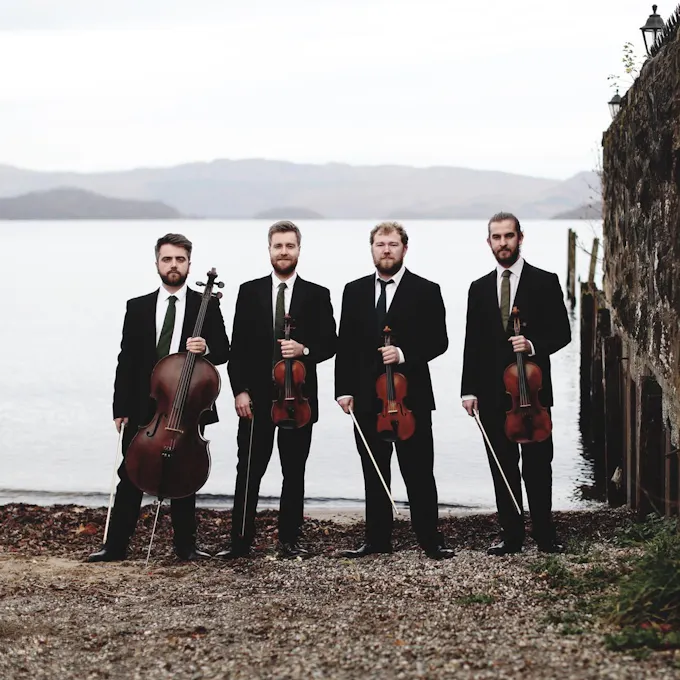 The musicians of Maxwell Quartet holding their instruments next to a stone wall, the sea behind them