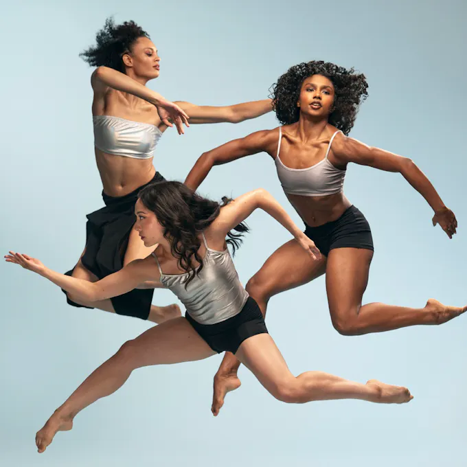 Three female dancers wearing black shorts and silver tops leap into the air