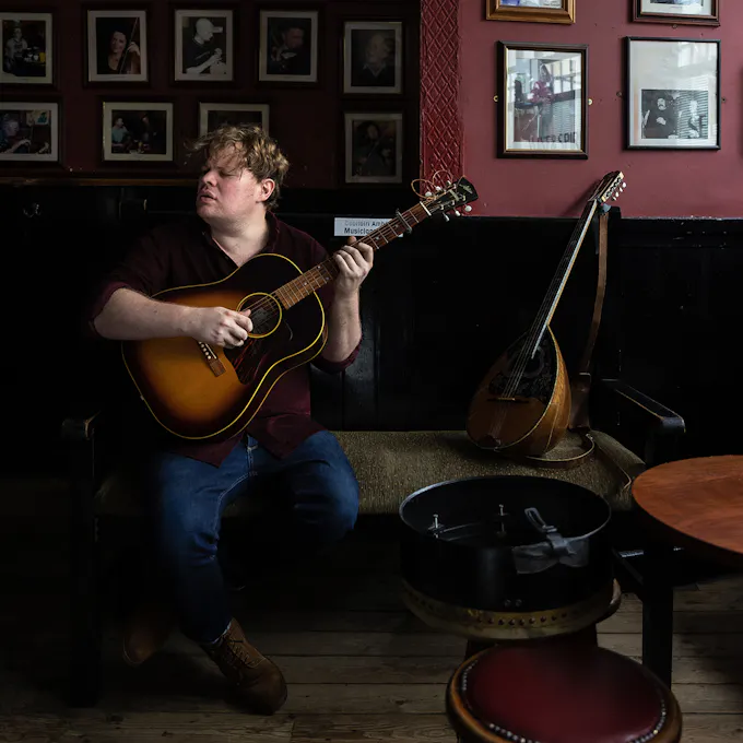 Macdara Yeates plays guitar in a pub, a wall of photos behind him