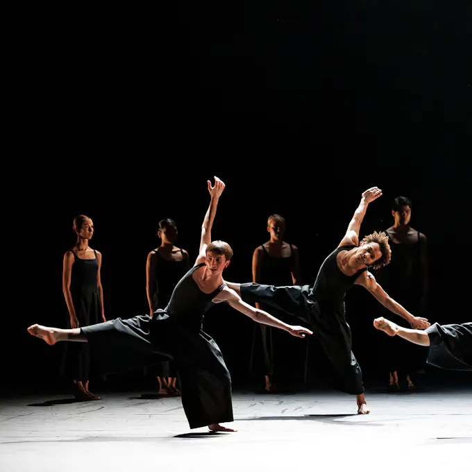 Two male dancers perform with arms and legs outstretched, four dancers standing behind them