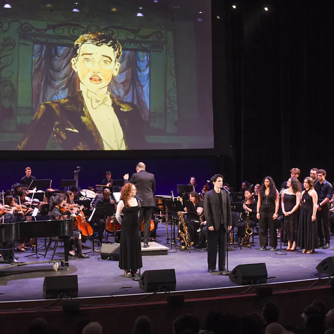 A choir, male and female singer, and orchestra perform on stage in front of a screen projecting an illustration of a young man wearing a tuxedo