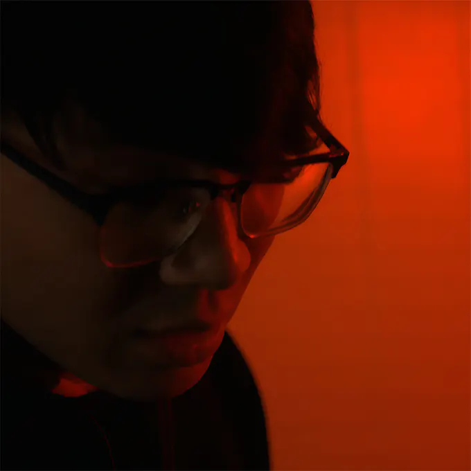 A young man wearing glasses, head bowed, against an orange backdrop