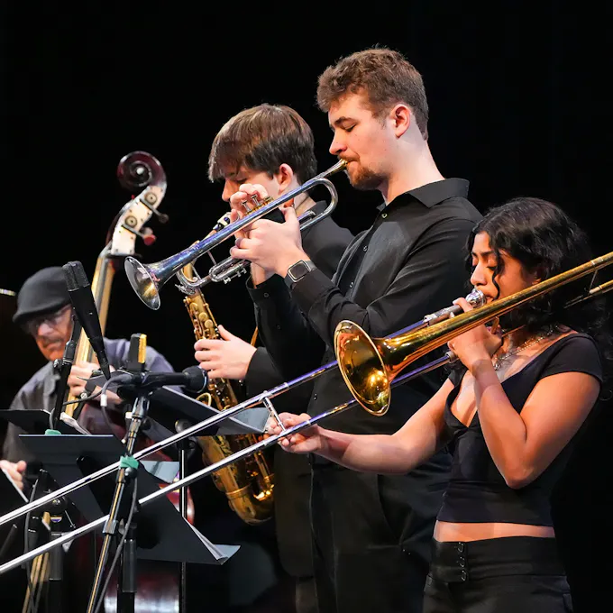 A young woman plays trombone and two young men play trumpet and saxophone as another man plays string bass