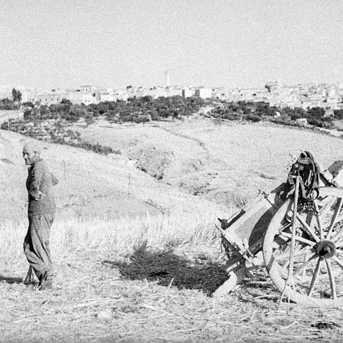 Grainy black & white photo of a man standing in a field across from a wagon, a city in the distance