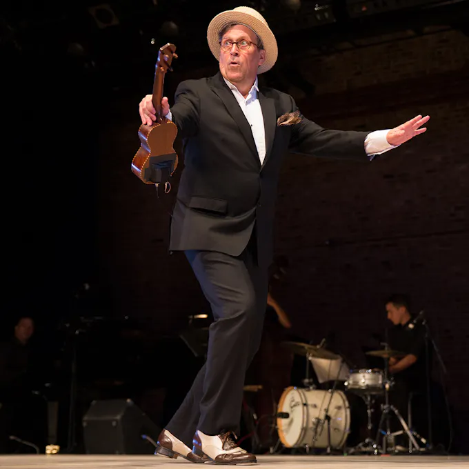 A man in a suit and straw hat holding a ukulele tap dances on stage as a drummer plays behind him
