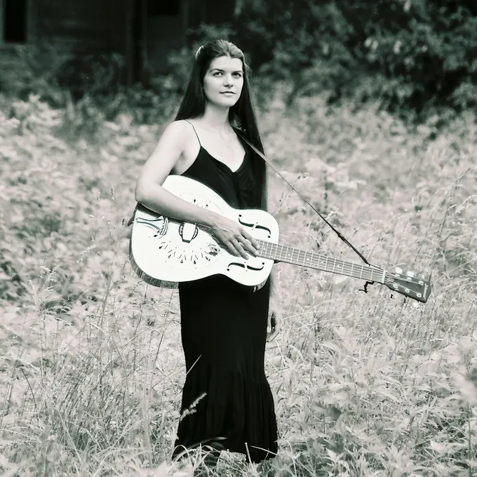 A young woman wearing a black dress stands in a field holding a guitar