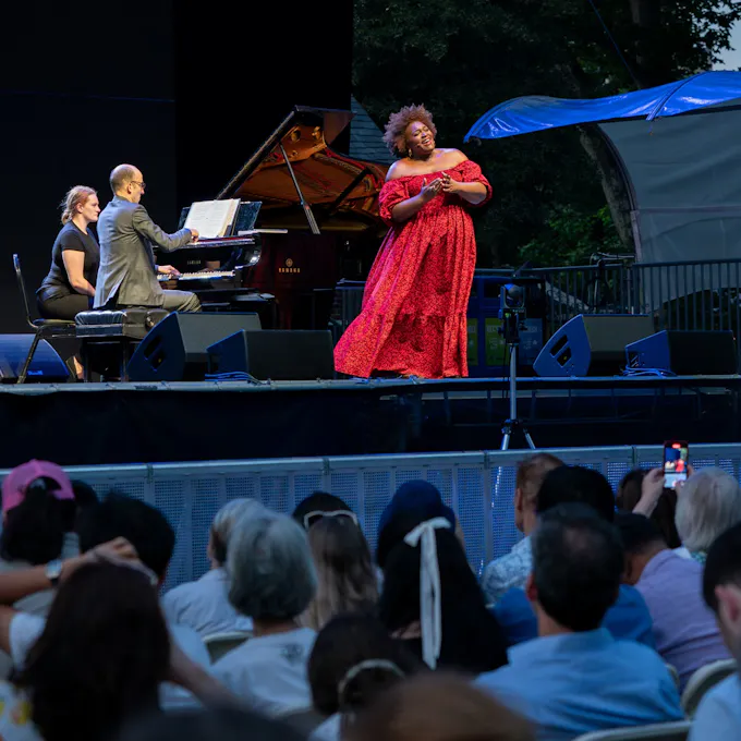 A woman wearing a flowing red gown sings on stage before an audience as a man plays piano behind her