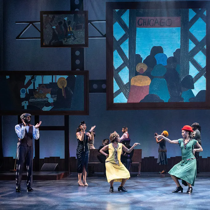 Two young women wearing flapper type dresses dance on stage as a man and woman in period costumes clap