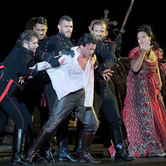 Four actors wearing period soldier uniforms hold back a man wearing black leather pants and white shirt