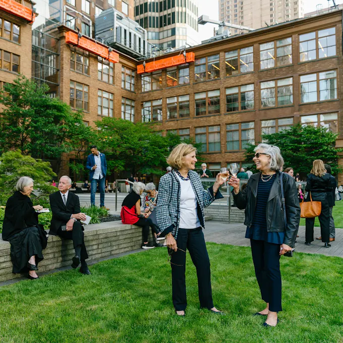 Two smiling women clink glasses on a rooftop terrace as others mingle behind them